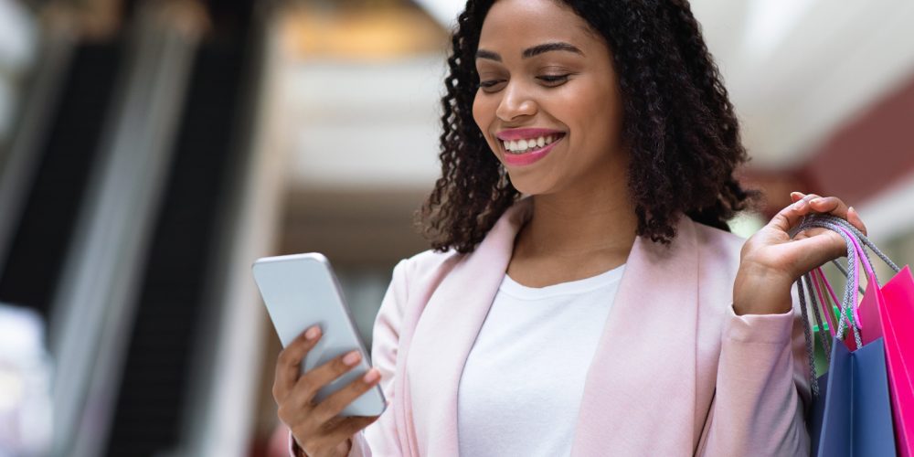 Discount App. Smiling African American Woman With Shopping Bags Using Smartphone In Mall, Checking Sale Offers, Free Space